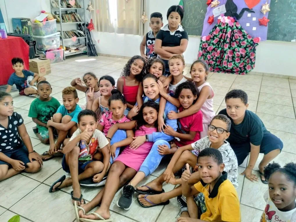 Grupo de crianças sorrindo, sentadas no chão de uma sala do Centro ao redor de uma educadora